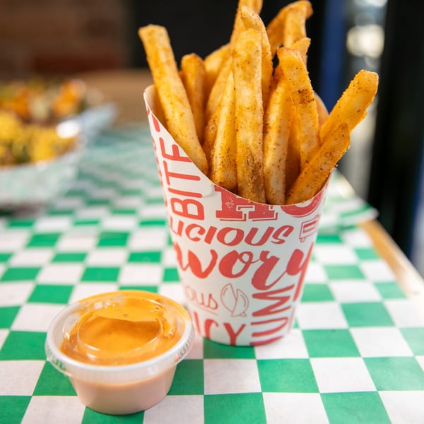 Seasoned fries in a paper cup with a side of dipping sauce on a green and white checkered table.