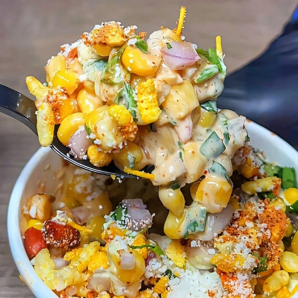 A spoonful of Nishaan's corn salad with onions, spices, and creamy dressing, lifted from a white bowl.