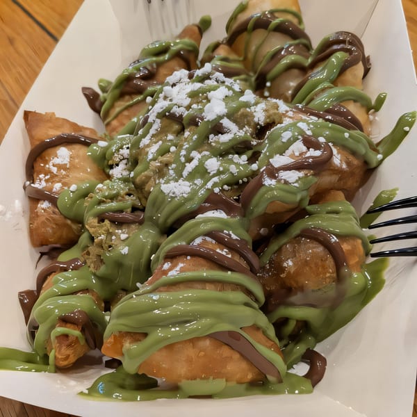 Fried pastries drizzled with green and brown sauces, topped with powdered sugar in a white container.